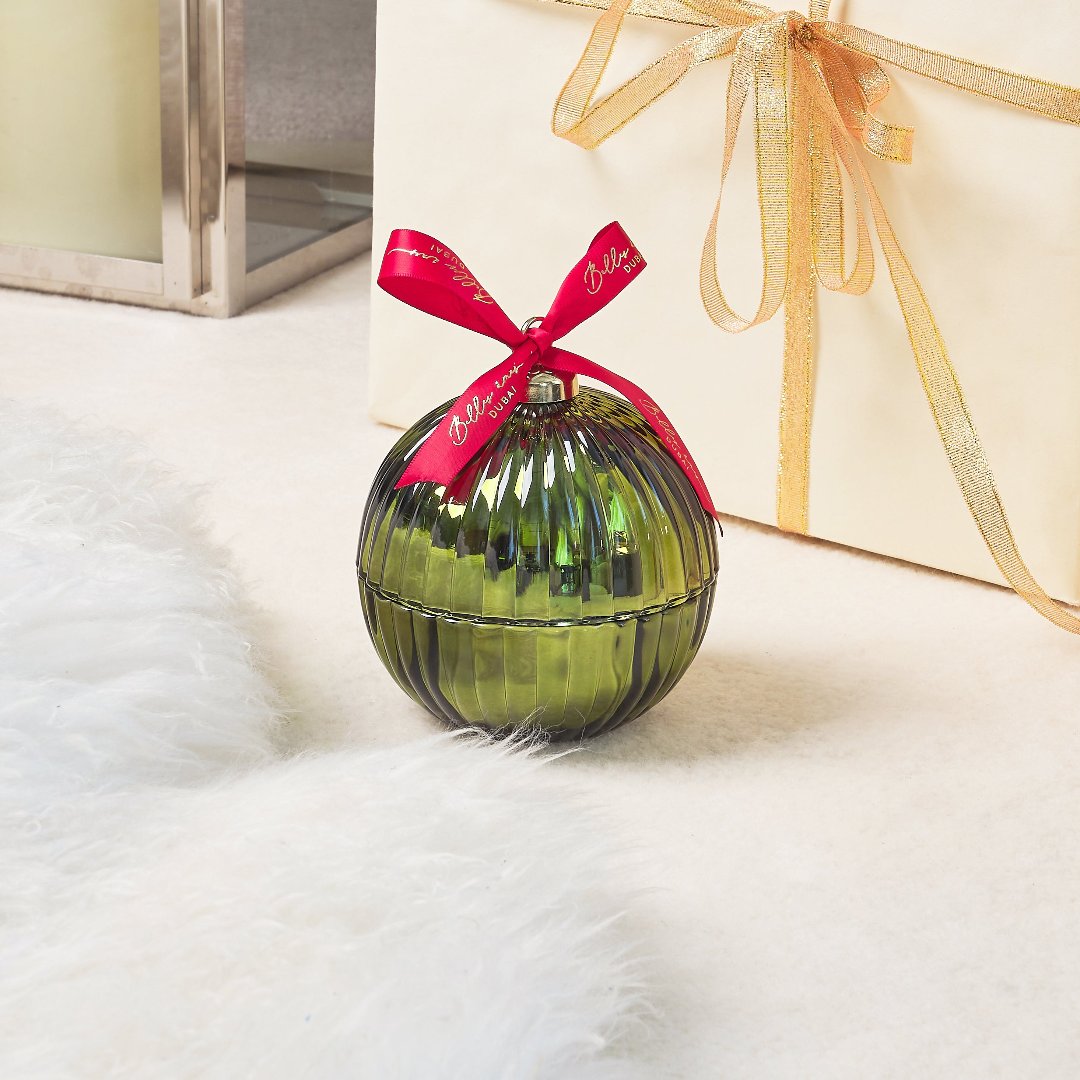 green bauble with red ribbon on a white fur rug and behind it a white gift box with some gold ribbon